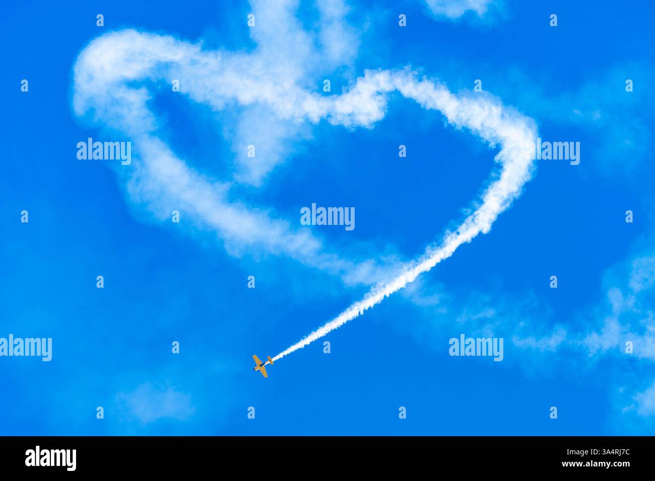 Ein kleines Kunstflugzeug hinterlässt eine weiße Rauchspur, die eine Herzform am blauen Himmel bildet. Stockfoto