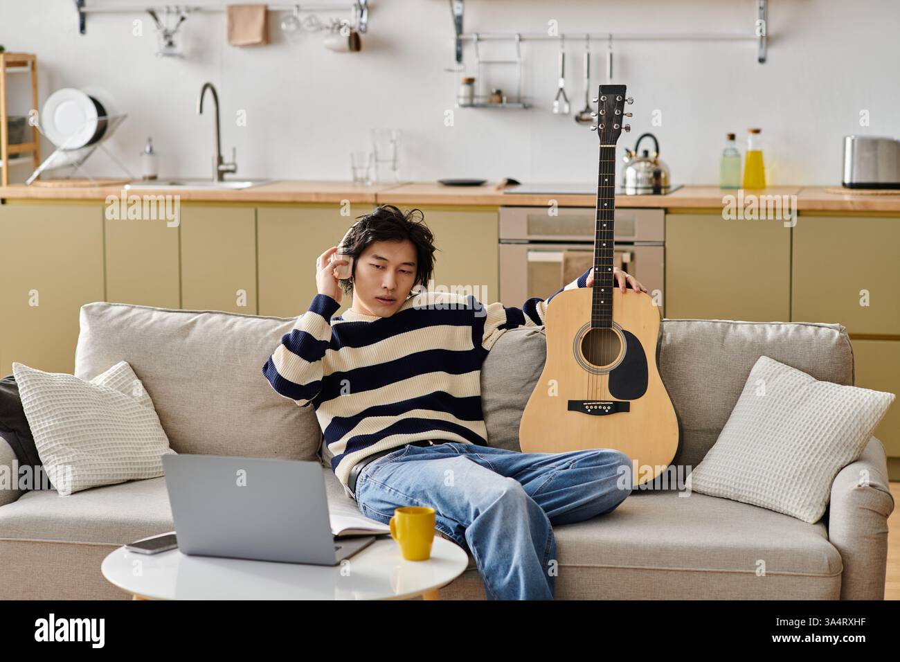 Ein hübscher junger asiatischer Mann sitzt bequem auf einer Couch, während er Gitarre übt, konzentriert und inspiriert. Stockfoto