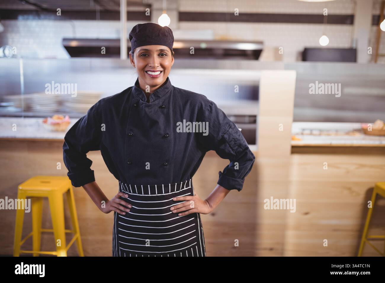 Koch in Uniform lächelt selbstbewusst in moderner Café-Küche mit Händen auf den Hüften Stockfoto