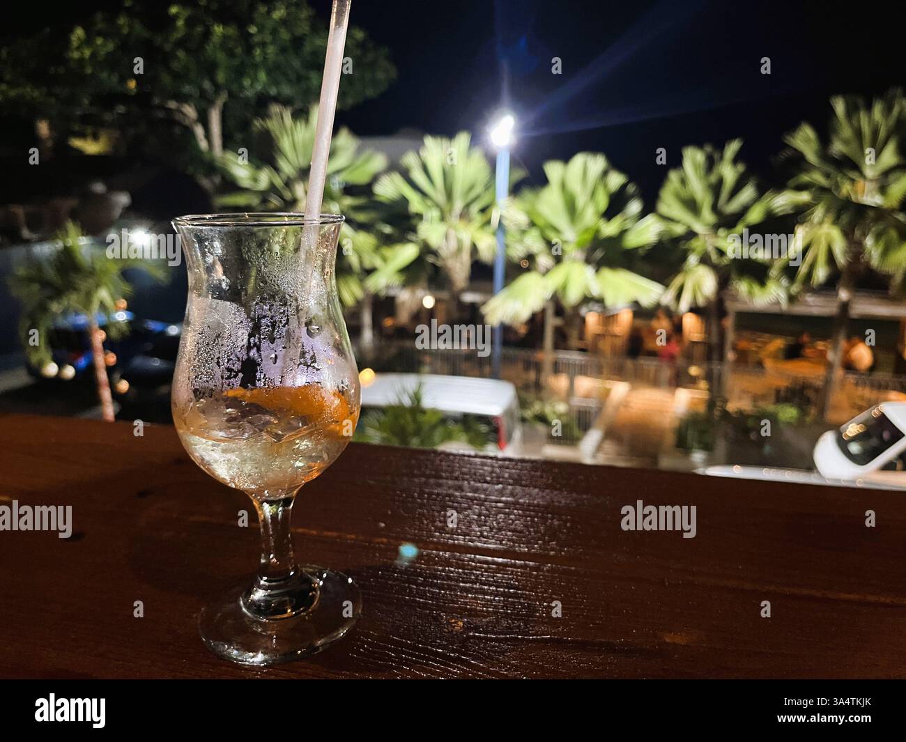 Ein leeres Cocktailglas, bedeckt mit Kondenswasser, liegt auf dem Geländer einer Dachterrasse mit Blick auf eine Palmenreihe auf Barbados. Stockfoto