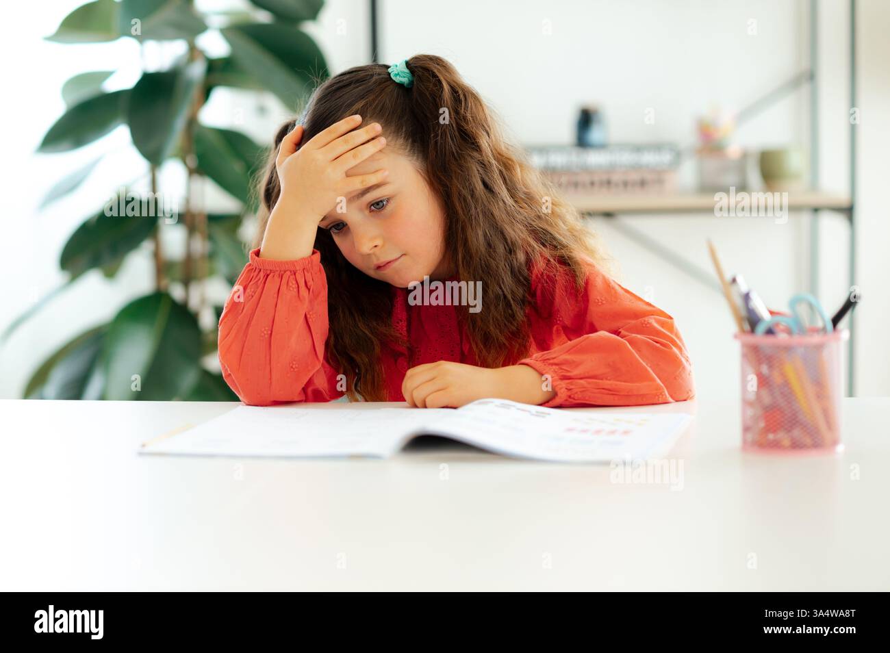 Müde gelangweilte kaukasische Schulmädchen in roter Bluse sitzt an einem weißen Schreibtisch und legt ihre Stirn auf der Hand, während sie ihr Arbeitsbuch mit frustriertem Ausdruck während der Hausaufgaben zu Hause ansieht Stockfoto