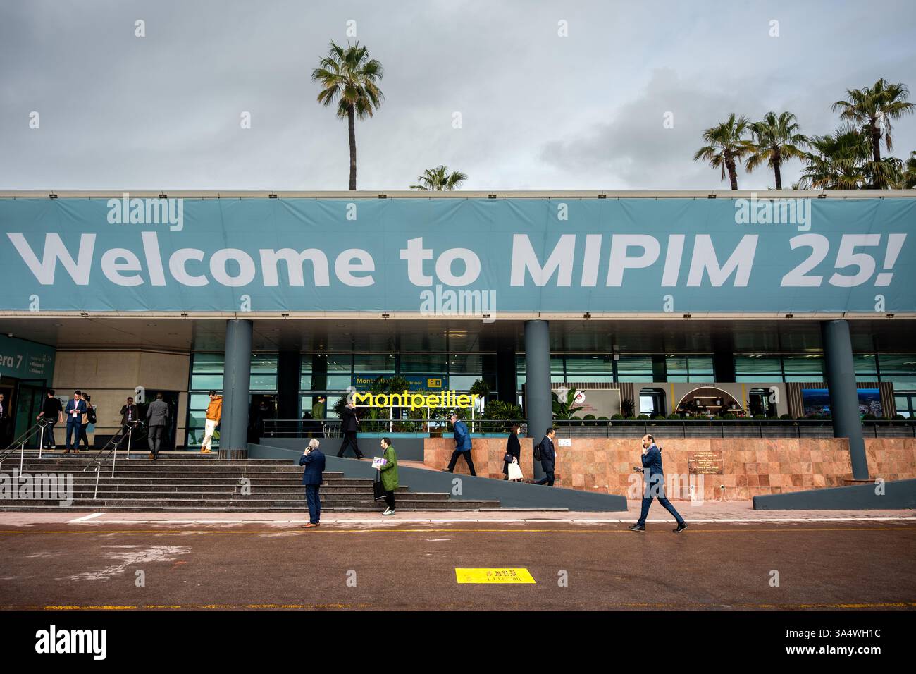Cannes, Frankreich. März 2025. Männer und Frauen passieren den Eingang zur internationalen Messe MIPIM mit einem Banner mit der Aufschrift „Willkommen bei MIPIM (Marché International des Professionnels de l'Immobilier). Das MIPIM (Marché International des Professionnels de l'Immobilier) bringt Fachleute der Branche (Bauunternehmer, Architekten, Immobilienmakler) im Palais des Festivals zusammen. (Foto: Laurent Coust/SOPA Images/SIPA USA) Credit: SIPA US/Alamy Live News Stockfoto