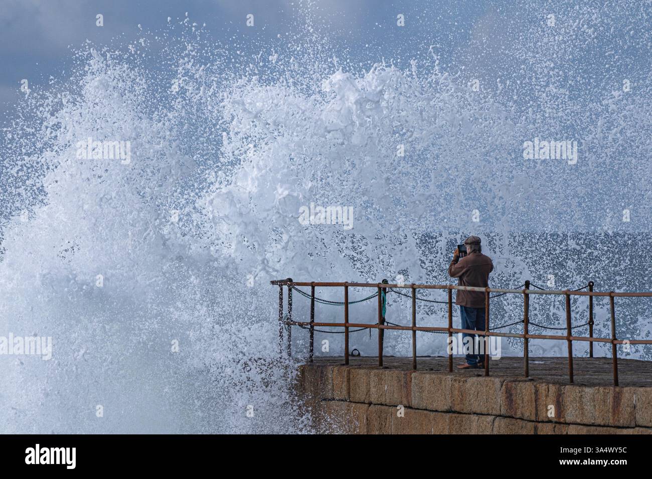 Ein Beobachter auf dem Pier in Porthleven wird fast nass, als eine Welle vor ihm bricht und überall Sprühnebel auftaucht. Stockfoto