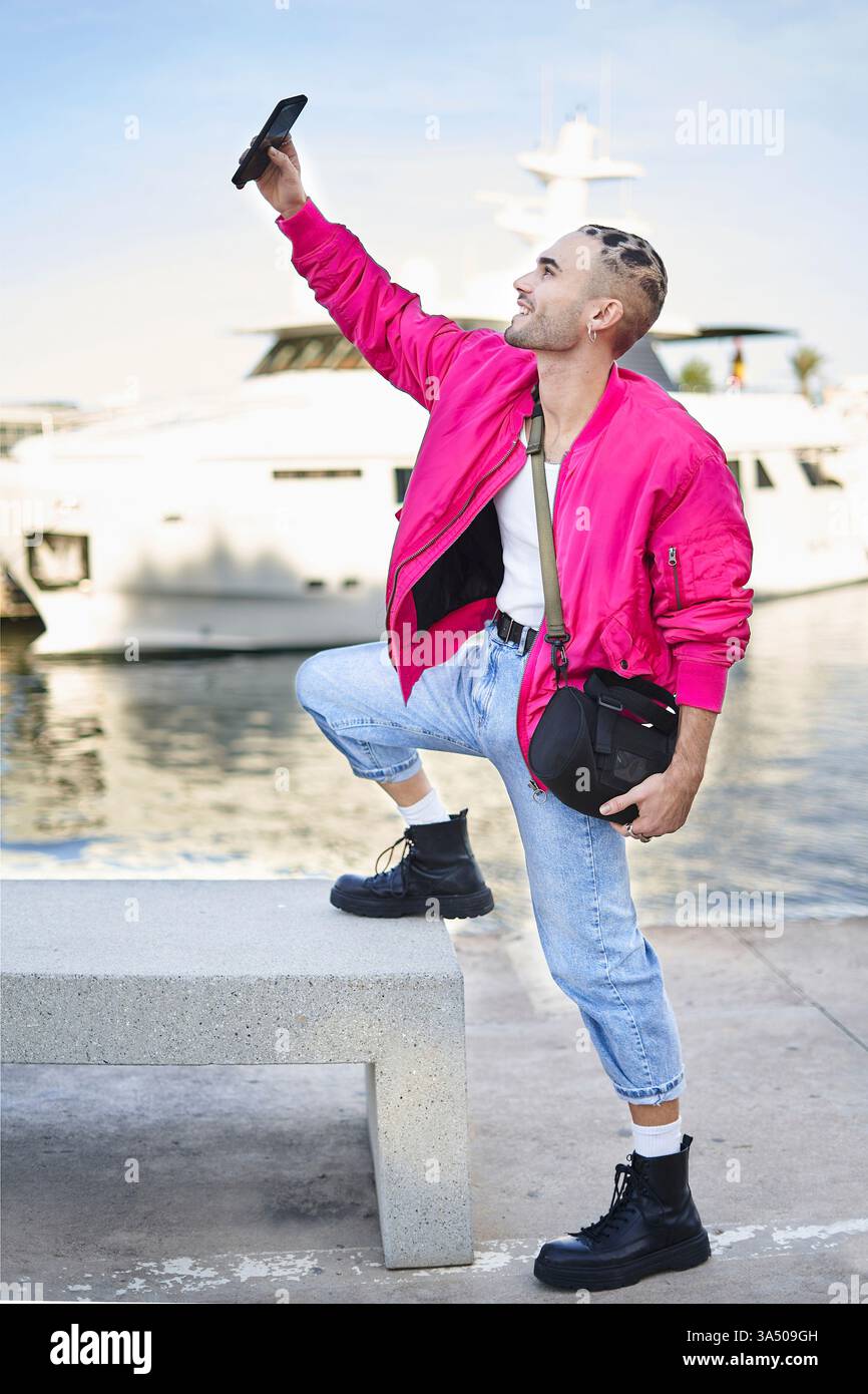 Ein lächelnder hispanischer Mann macht ein Selfie im Freien und steht mit einem Fuß auf einer Bank am Yachthafen. Der ungezwungene, unbeschwerte Moment sorgt für eine moderne Social-Media-Atmosphäre auf Reisen und am Wasser. Ideal für Lifestyle-, Tourismus- und Branding-Projekte. Stockfoto