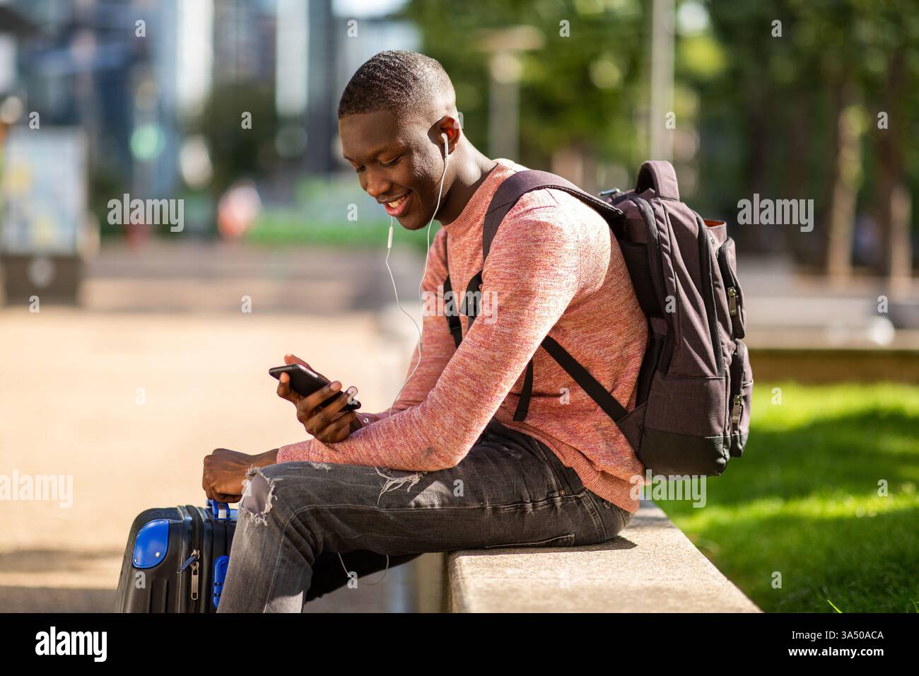 Porträt eines lächelnden jungen afroamerikanischen Mannes draußen, der mit einem Koffer sitzt und auf sein Handy schaut. Diese reisebereite Szene fängt den urbanen Lebensstil und die Vorfreude auf den Beginn der Reise ein. Ideal für Reisen, Tourismus und Geschäftsreisen. Stockfoto