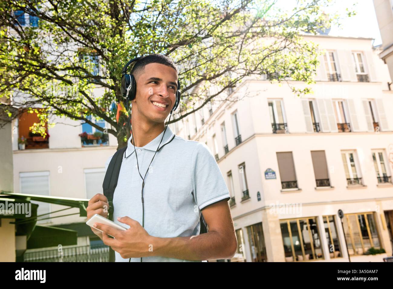 Ein arabischer Mann steht tagsüber auf einer Stadtstraße, hält ein Handy und schaut ihm über die Schulter. Diese urbane Straßenszene vermittelt einen modernen, vernetzten Lebensstil mit Kopfhörern und einem Rucksack, der auf Reisen und Freizeit hinweist. Ideal für Technologie, mobile Konnektivität oder multikulturelle urbane Kampagnen. Stockfoto