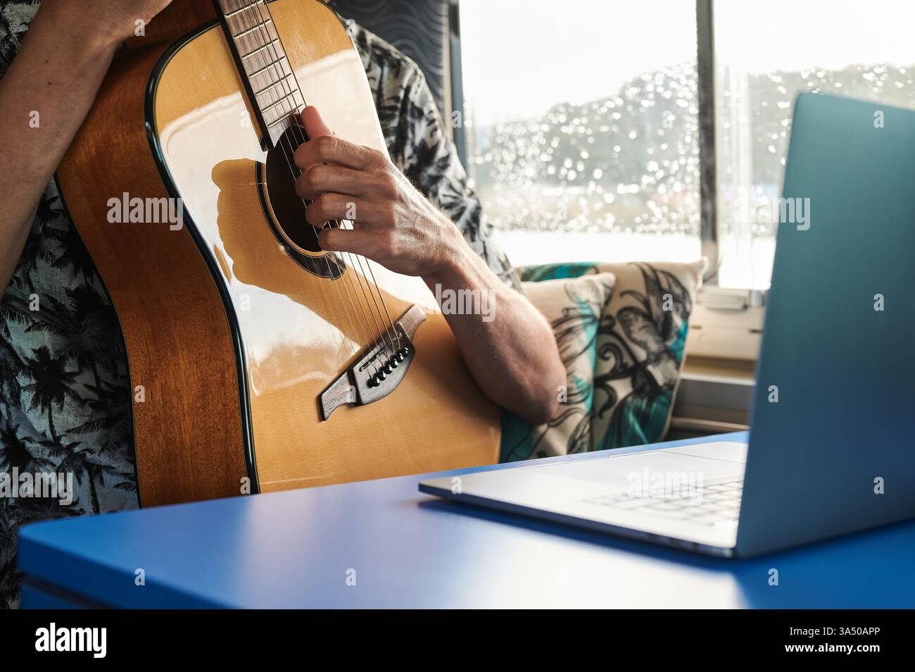 Crop zeigt einen gesichtslosen männlichen Musiker, der eine akustische Gitarre spielt, während er eine Videostunde auf einem Laptop in einem Van ansieht. Eine einzigartige Mischung aus Musikpraxis und digitalem Lernen für mobile oder Reisekontexte. Ideal für Inhalte über Online-Gitarrenunterricht, Fernmusiker und Fortbildung unterwegs. Stockfoto