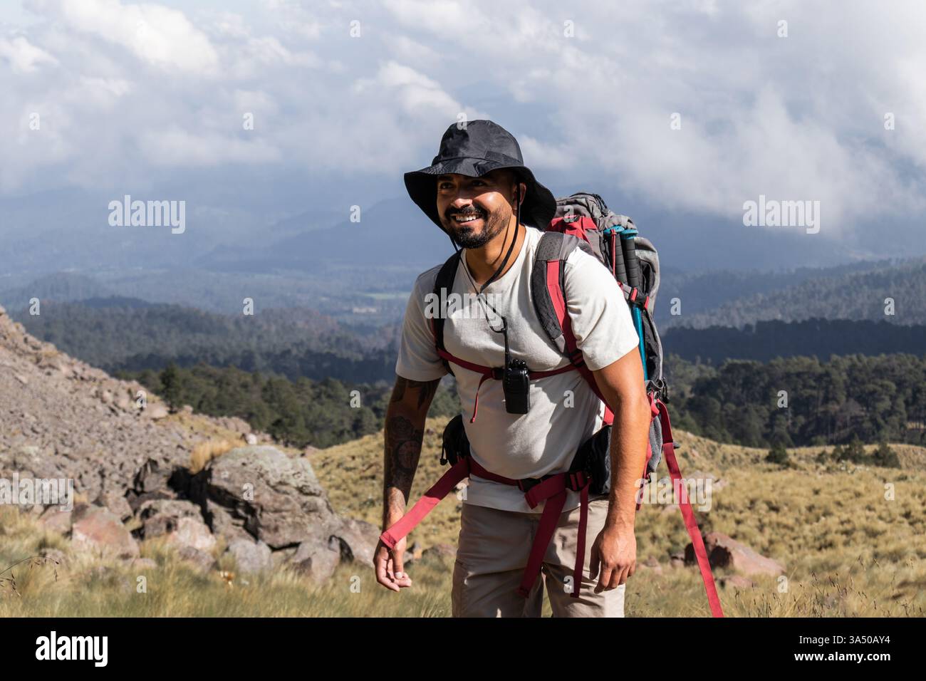 Fröhlicher männlicher Wanderer mit Bart und Tätowierungen mit Sonnenhut und Rucksack steht auf einem Bergweg. Er blickt mit einem breiten Lächeln direkt in die Kamera. Dieses Outdoor-Abenteuer vor einer vulkanischen Landschaft eignet sich ideal für Reise- und Wandermotive. Stockfoto