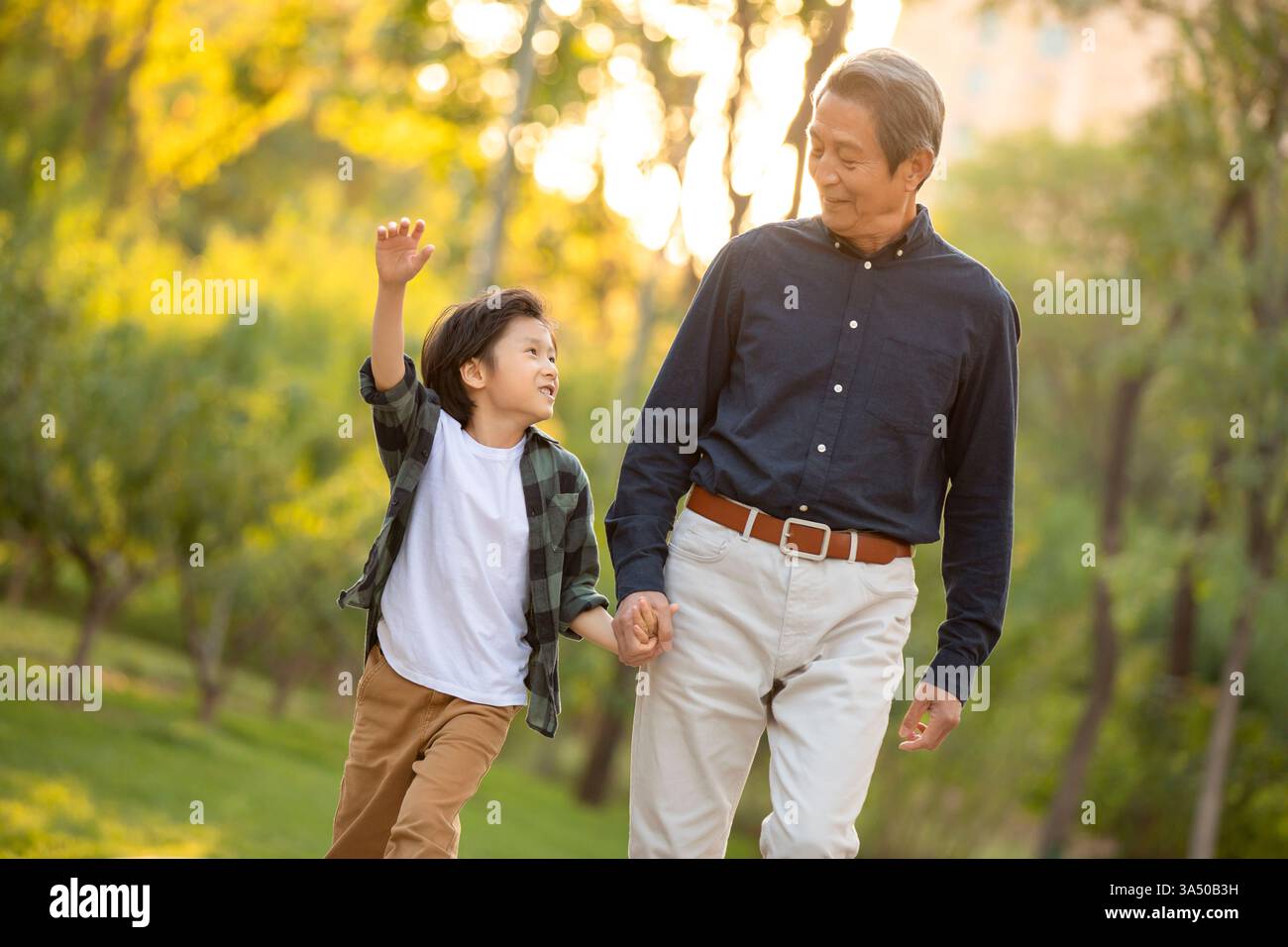 Chinesischer Großvater und Enkel, die Hände halten, gehen bei Sonnenaufgang zusammen im Park Stockfoto
