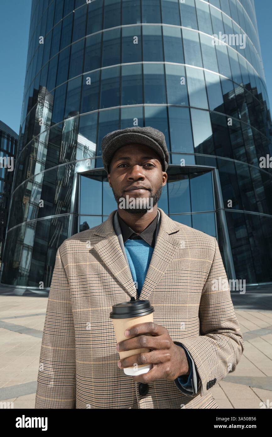 Porträt eines Afrikaners in einem Anzug, der draußen in der Stadt steht und die Kamera ansieht, während er Kaffee trinkt. Ein poliertes, professionelles Stadtporträt, das sich für Corporate Branding und Visuals des Stadtlebens eignet. Auffälliger, moderner Look für Business- und Urban-Kampagnen. Stockfoto