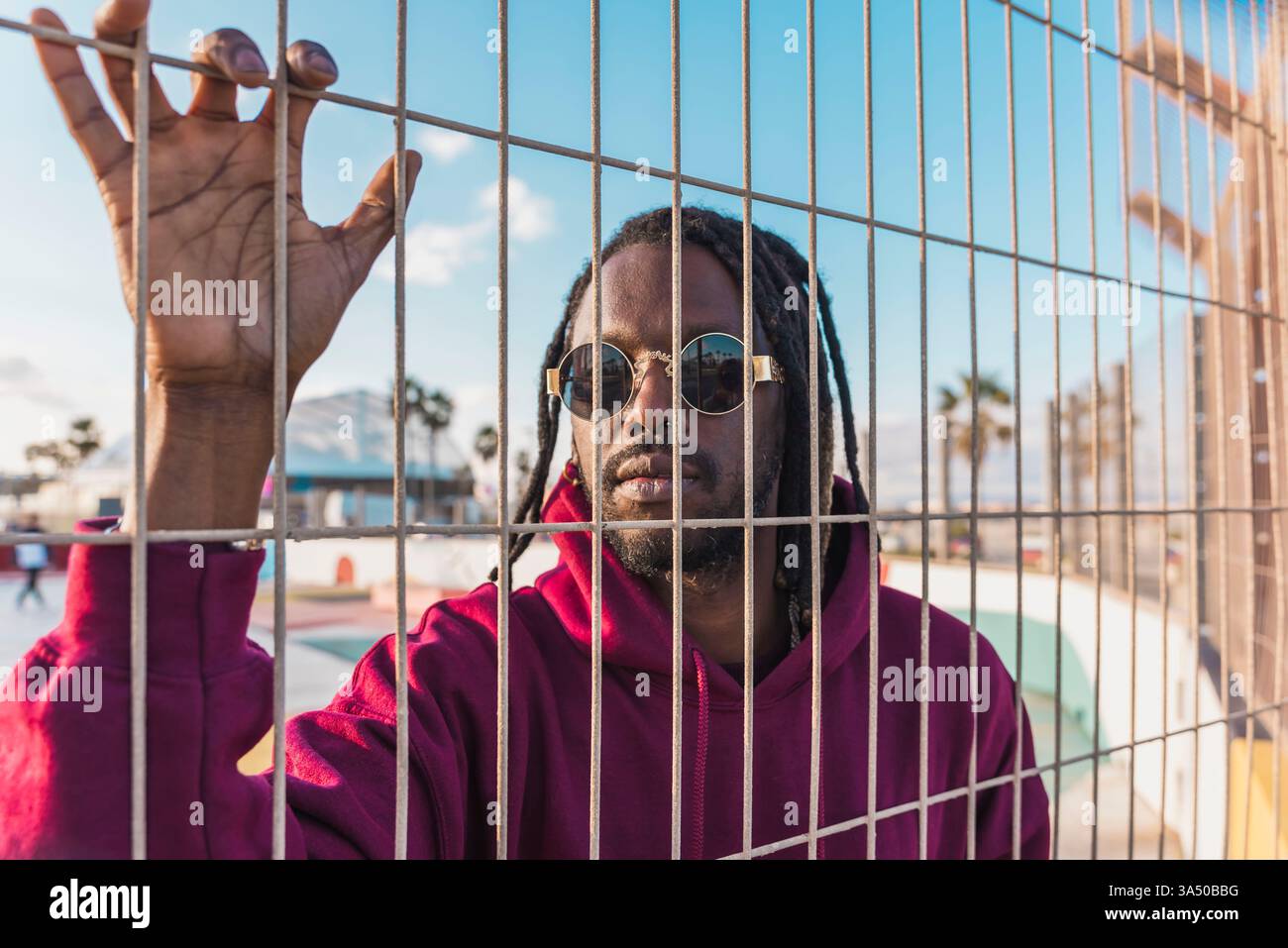 Serious Black man mit Dreadlocks, Sonnenbrille und Hoodie, steht an einem sonnigen Tag hinter einem Metallzaun in einem Park. Der urbane Streetstyle-Look kombiniert Freizeitkleidung mit durchdachter Stimmung und ist ideal für Lifestyle und urbane Kampagnen. Stockfoto