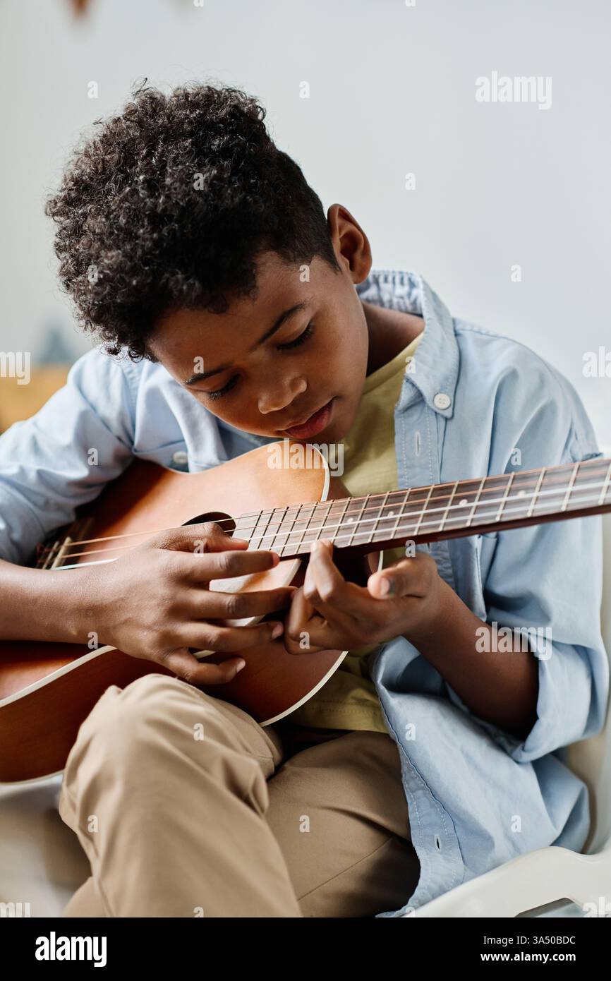Serious Black Boy saß im Kreuz auf einem Stuhl und spielte zu Hause Gitarre. Ein pädagogisches und musikalisches Motiv, das sich für Unterricht, Praxis und Talentförderungskampagnen eignet. Das Bild vermittelt Fokus, Disziplin und Kreativität im häuslichen Umfeld. Stockfoto
