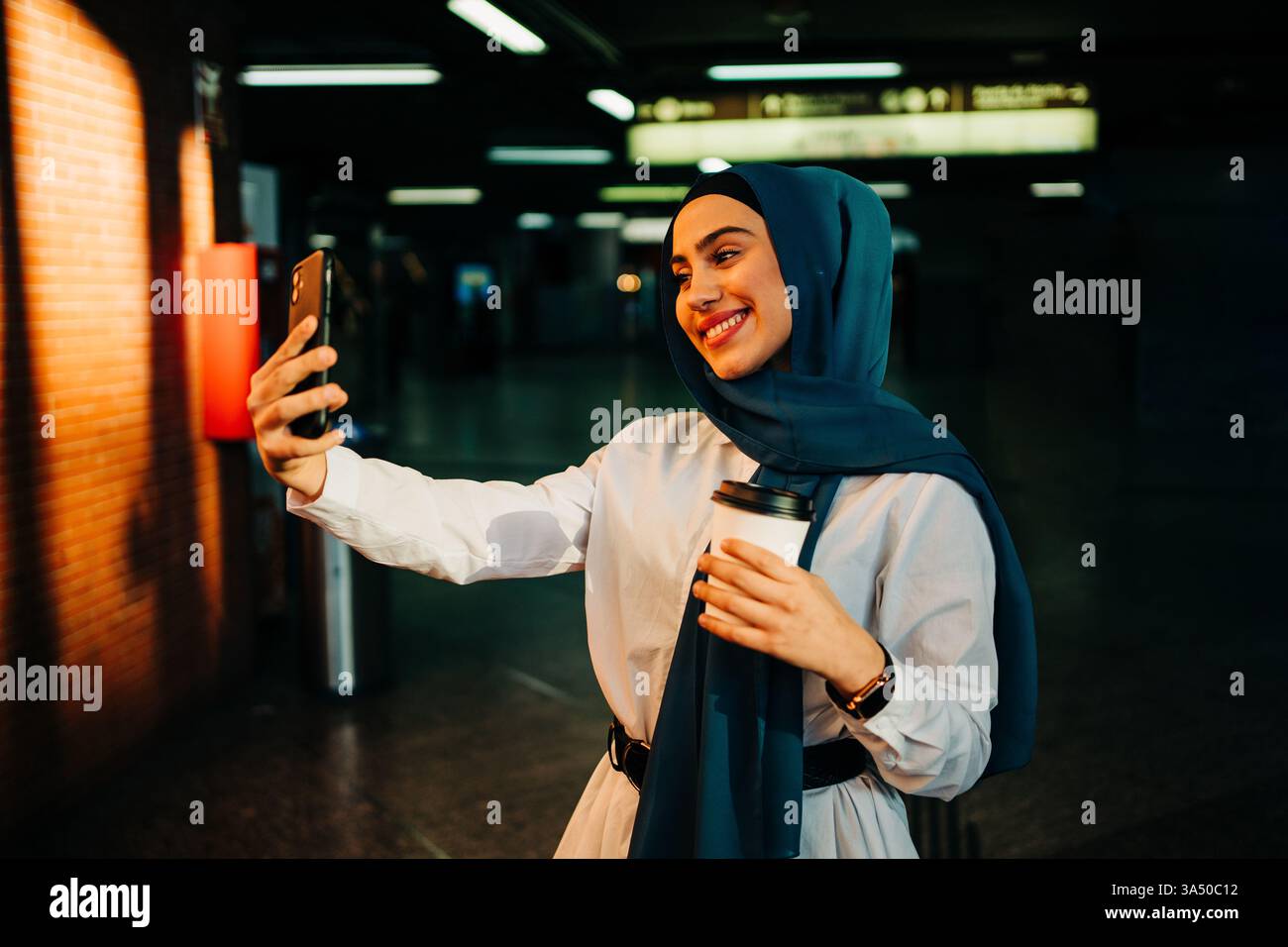 Eine Frau aus dem Nahen Osten, die einen Hijab trägt, macht ein Selfie am Bahnhof. Dieser urbane Moment der Konnektivität lässt Reisen und Social Sharing vermuten. Stockfoto