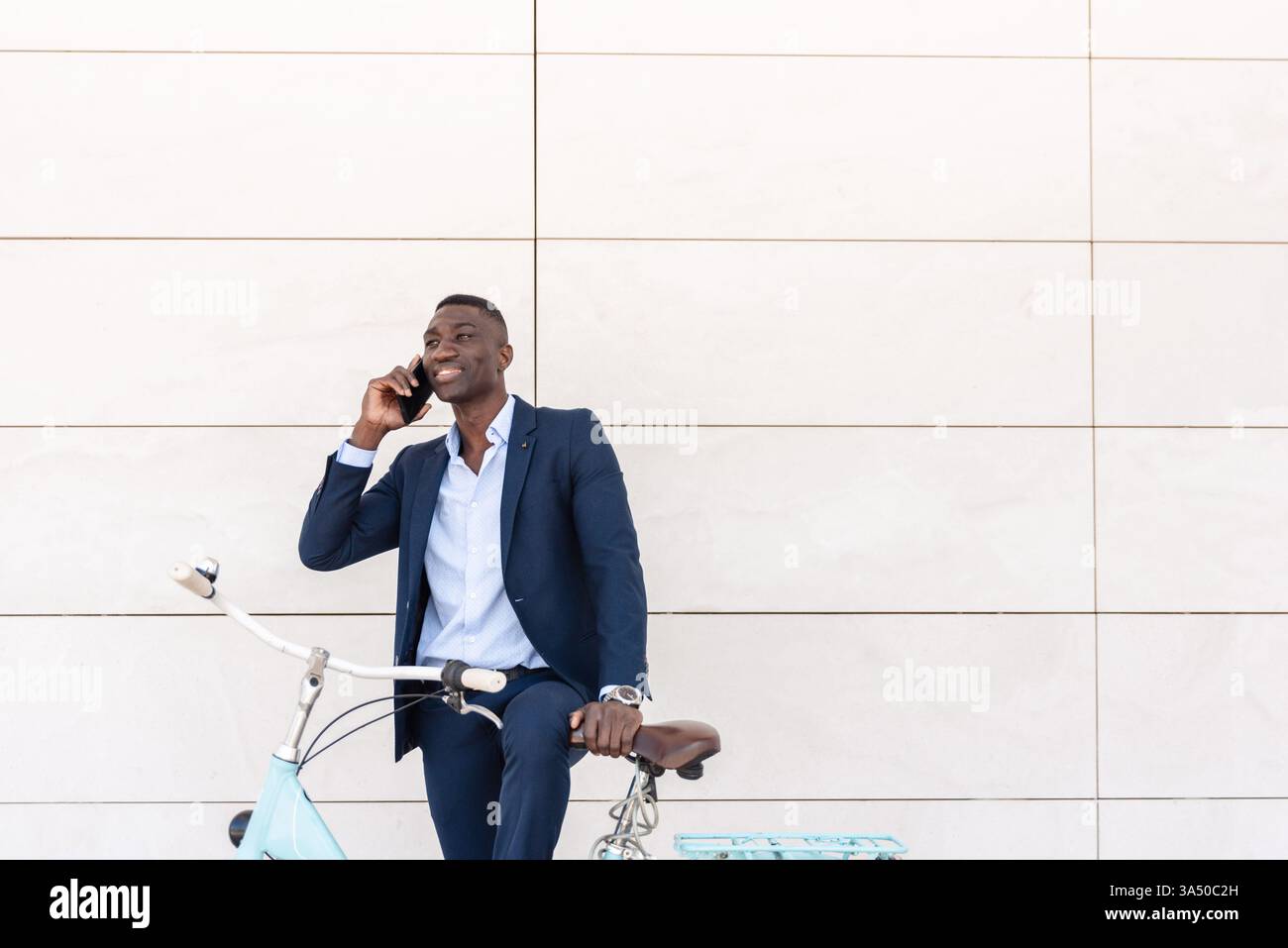 Ein junger afroamerikanischer Geschäftsmann in formeller Kleidung lächelt, während er auf einem Handy spricht und sich auf einem Fahrrad auf einer städtischen Straße stützt. Geeignet für Unternehmens-, Start-up- und urbane Lifestyle-Kampagnen, die Mobilität und Konnektivität vermitteln. Ideal für Geschäftskommunikation, Netzwerke und mobile Bürokonzepte. Stockfoto
