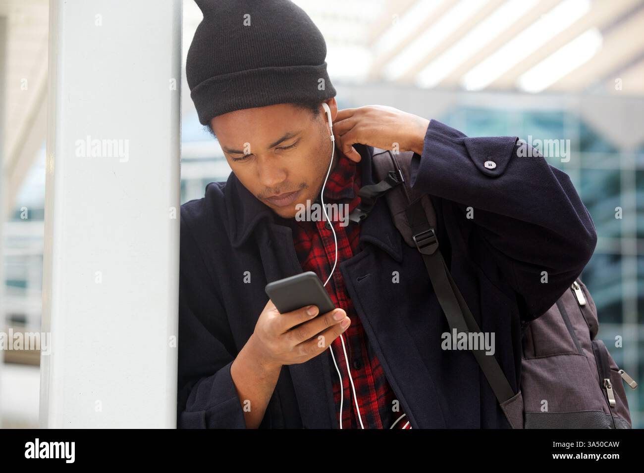 Moderner Reisender in einem Flughafenterminal mit Smartphone mit Ohrhörern. Ein Schwarzer in seinen 30ern, stilvoll und fokussiert, verkörpert einen urbanen, technisch freundlichen Lebensstil. Stockfoto