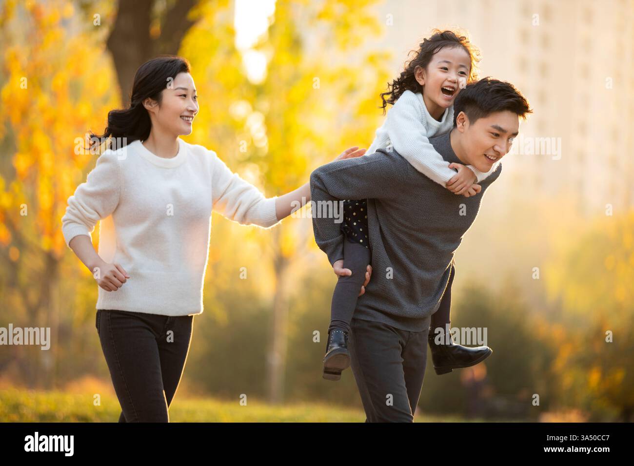 Fröhliche chinesische Familie, die an einem sonnigen Tag einen Huckepack-Ritt in einem Park genießt. Dieser warme Moment im Freien unterstreicht Zweisamkeit und zwanglose Erziehung – perfekt für den Familienstil oder die Erziehung. Ideal für Artikel über Wochenendaktivitäten, Herbsttage und multikulturelle Familien. Stockfoto