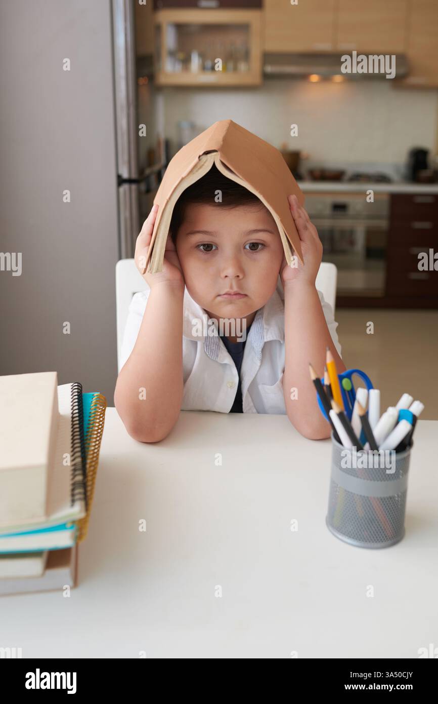 Der Mixed Race Boy hält ein offenes Buch über den Kopf, während er zu Hause an einem Esstisch sitzt. Dieser ehrliche Lernmoment unterstützt das Lernen zu Hause, das Lesen und die Bildung von Bildern mit verschiedenen Familien. Stockfoto