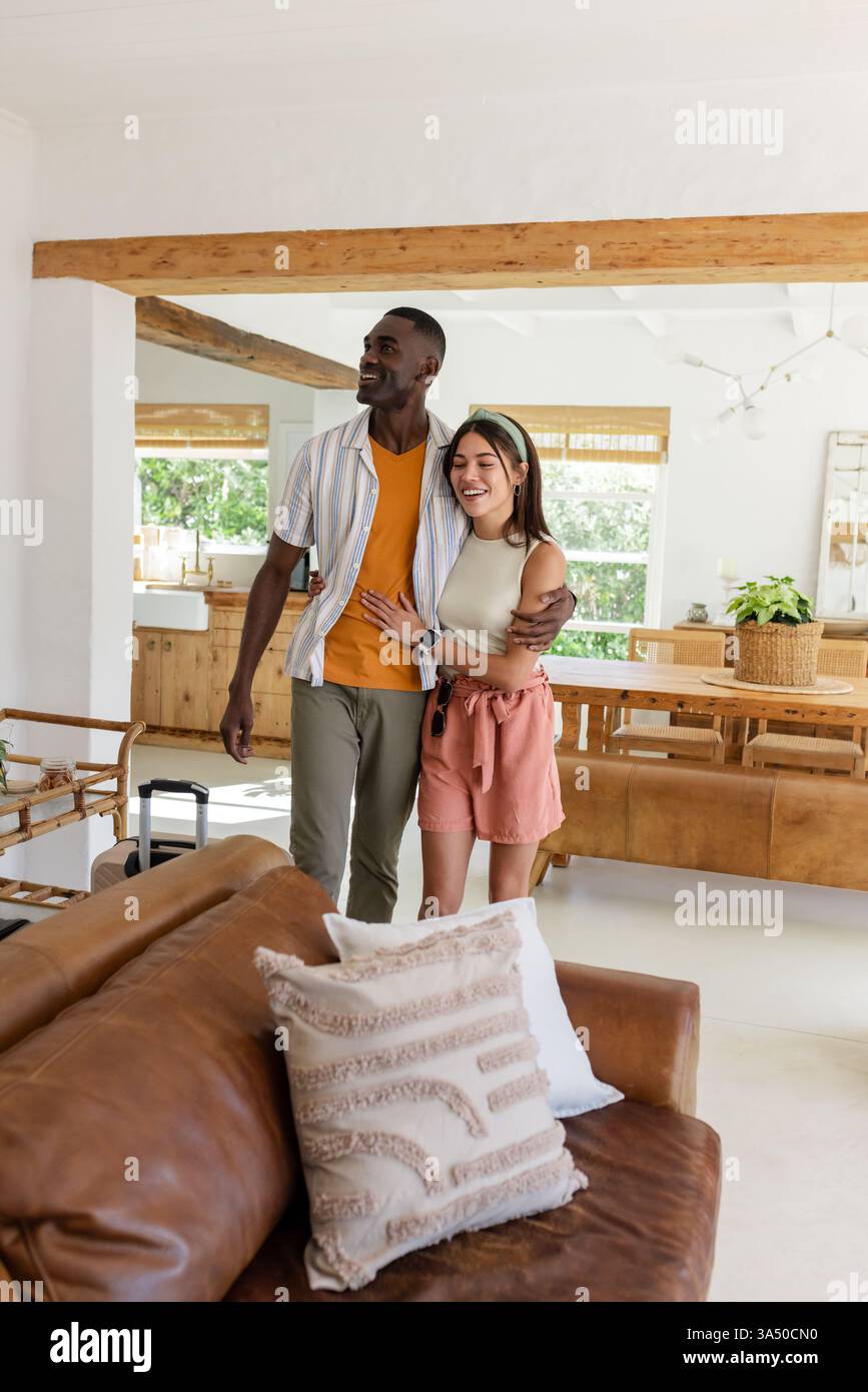 Glückliches vielseitiges Paar, das gemütliche Ferienhaus erkundet, sich umschließt und im Wohnzimmer lächelt Stockfoto