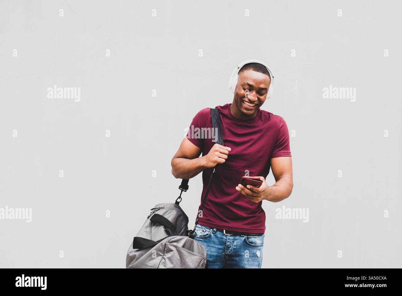 Porträt eines afroamerikanischen Mannes, der auf sein Handy schaut, während er eine Tasche trägt und Kopfhörer trägt. Eine urbane Lifestyle-Szene, in der mobile Technologien, Pendelfahrten und Musik unterwegs im Vordergrund stehen. Ideal für Inhalte über den täglichen Einsatz von Technologien, Reisen oder das Leben in der Stadt. Stockfoto