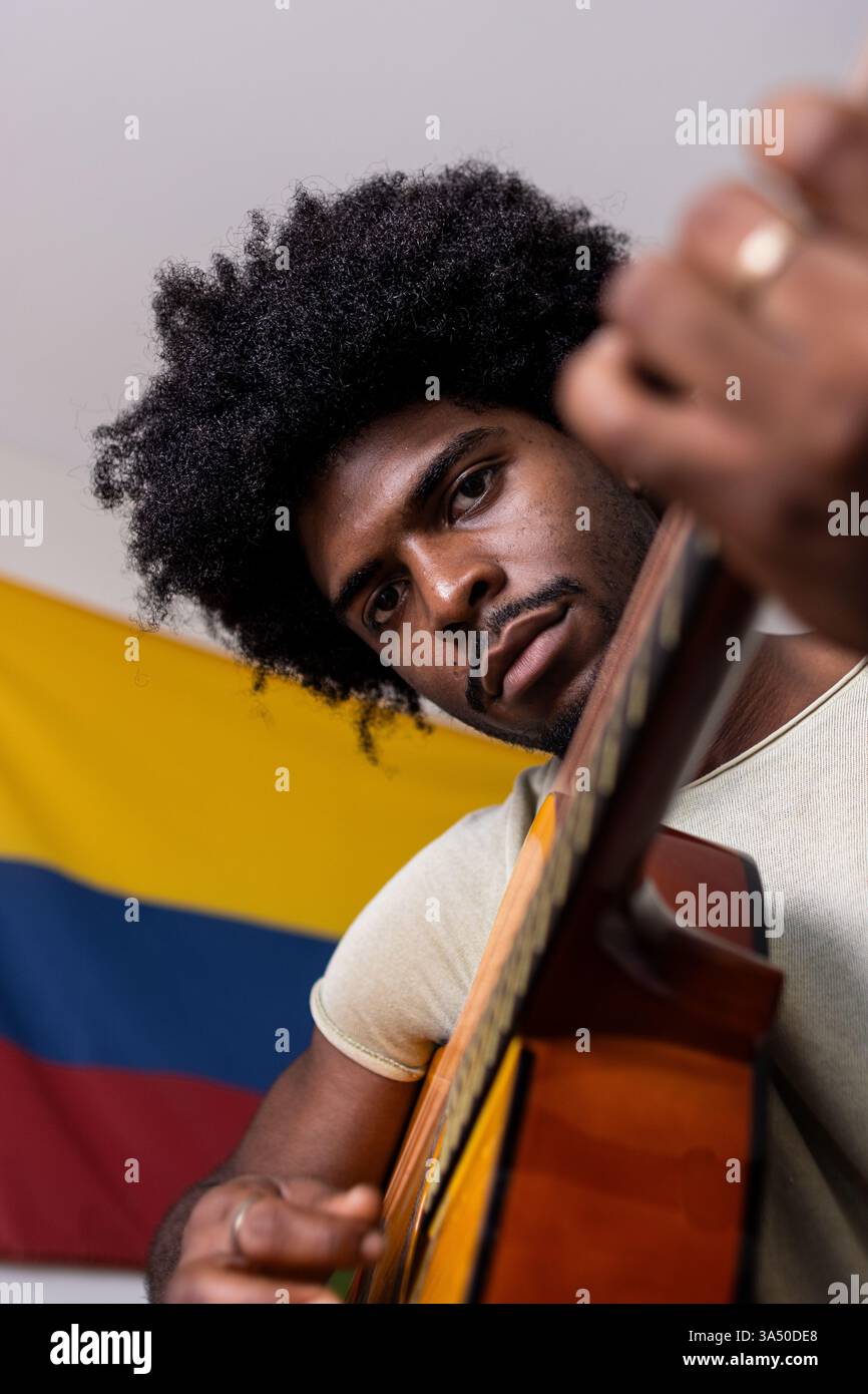 Porträt eines selbstbewussten jungen Mannes mit Afro-Haaren, der zu Hause Gitarre übt, gegen eine kolumbianische Flagge. Dieses Porträt fängt Fokus und musikalische Talente in einem persönlichen Raum ein. Geeignet für Inhalte über lateinamerikanische Kultur, Musikunterricht und Porträtfotografie. Stockfoto