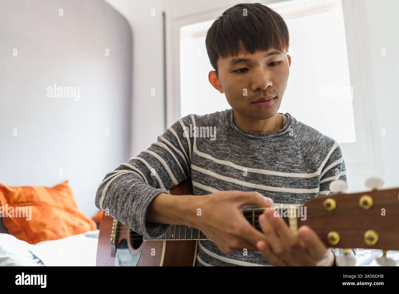 Ein asiatischer Mann sitzt zu Hause auf einem Bett und spielt Gitarre bei Tageslicht. Ein fokussierter, zwangloser Musiker, der drinnen übt, geeignet für Heimstudios oder Hobbyinhalte. Stockfoto