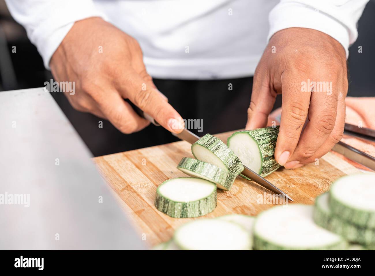 Anonymer männlicher Koch, der frische Zucchini auf einem Holzschneidebrett schneidet. Kulinarische Vorbereitungsszene konzentriert sich auf gesundes Kochen und Zutaten. Ideal für Rezepte, Ernährung und Visualisierungen in der Küche oder Lebensmittelindustrie. Stockfoto