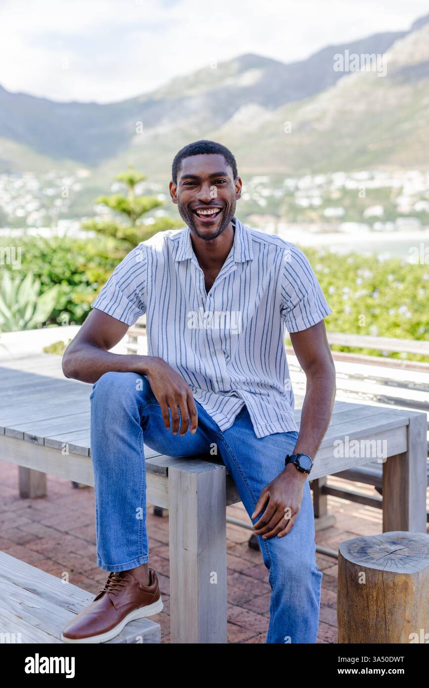 Lächelnder afroamerikanischer Mann, der auf einer Sitzbank sitzt und die Urlaubslandschaft genießt Stockfoto