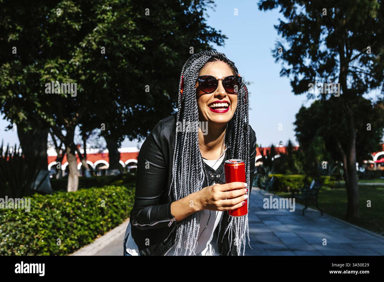 Fröhliche Hispanic Frau in einem Park an einem sonnigen Tag hält eine Bierdose und trägt Kopfhörer. Ihr lässiges Outfit und ihre Sonnenbrille spiegeln einen unbeschwerten urbanen Lebensstil und Freizeit im Freien wider. Ideal für Inhalte über den Sommer, das gesellschaftliche Leben und den Genuss von Getränken in öffentlichen Räumen. Stockfoto