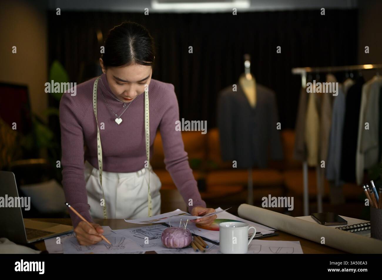 Die fokussierte Millennials asiatische Modedesignerin entwirft in ihrem Studio ein neues Kostümmodell. Umgeben von Stoffen und Designwerkzeugen konzentriert sie sich auf ihre Kollektion. Geeignet für Modedesign, Boutiquen-Branding und kreative Entrepreneurship-Visuals. Stockfoto