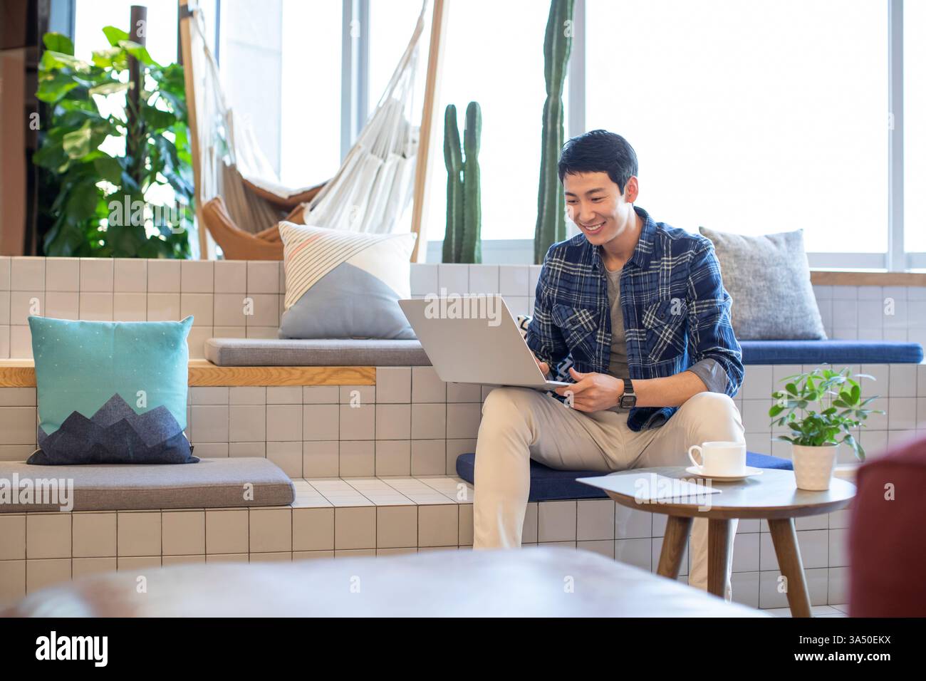 Ein lächelnder chinesischer Mann, der einen Laptop an einem Café-Tisch benutzt, um einen entspannten Lern- oder Arbeitszeitpunkt festzuhalten. Ideal für Funktionen im Universitätsleben, in der Campus-Kultur und im Online-Lernen. Ideal für Artikel über Technik, Fernarbeit oder Kaffeekultur in Asien. Stockfoto