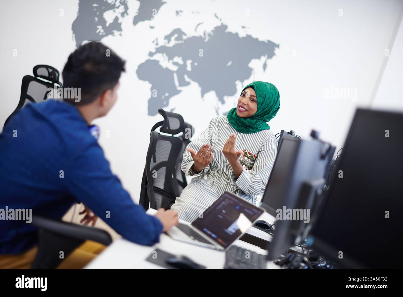 Schwarze Frau mit Hijab, die auf dem Schreibtisch sitzt und mit einem männlichen Kollegen aus dem Nahen Osten spricht, der am Tisch sitzt und im Büro einen Laptop hat Stockfoto