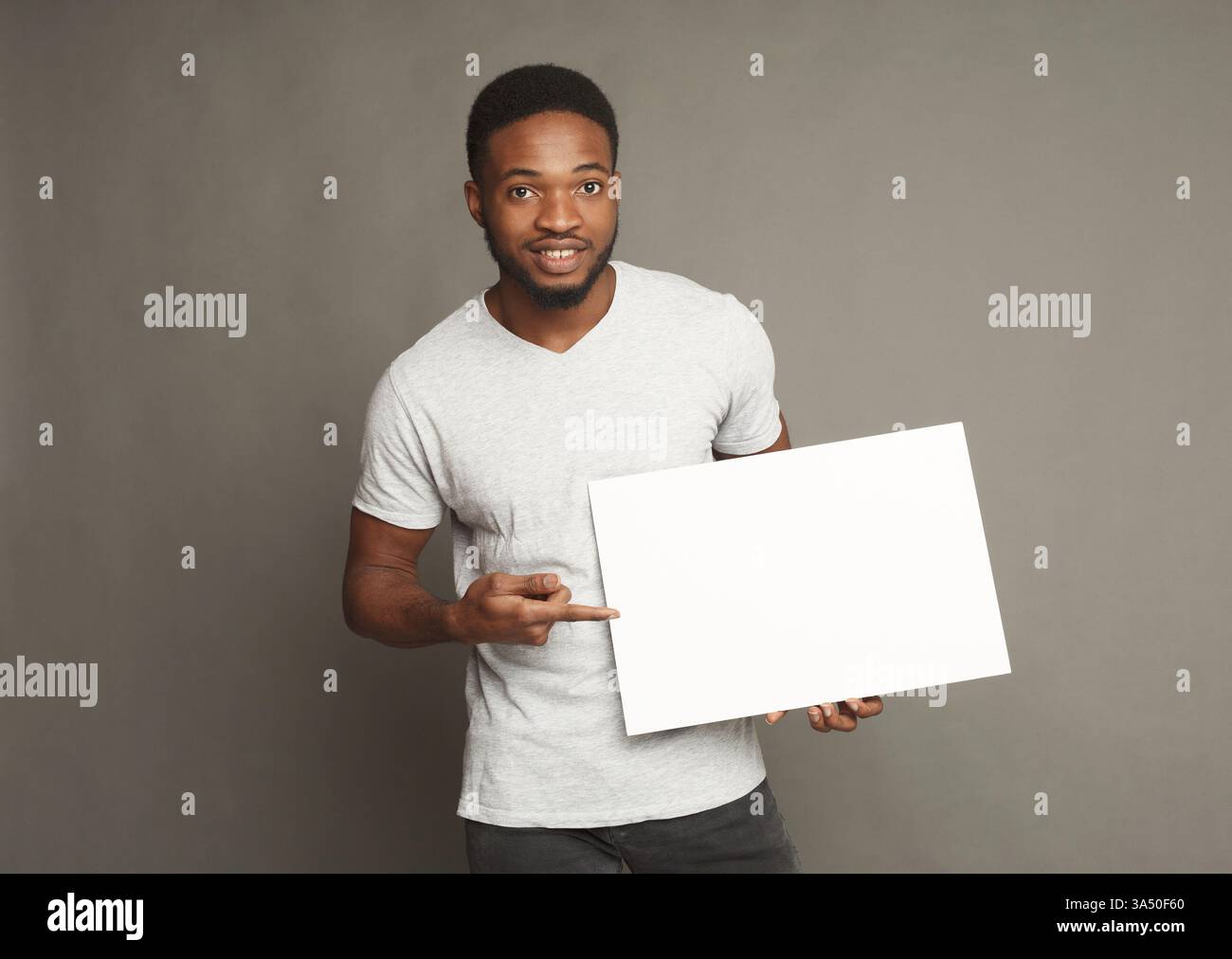 Ein lächelnder junger afroamerikanischer Mann hält ein leeres Whiteboard, ideal für Werbe- und Werbekampagnen. Er zeigt auf die Tafel vor einem neutralen grauen Hintergrund, wodurch er freien Kopierraum für Ihre Botschaft bietet. Perfekt für Marketinggrafiken, Banner und Produktaktionen. Stockfoto