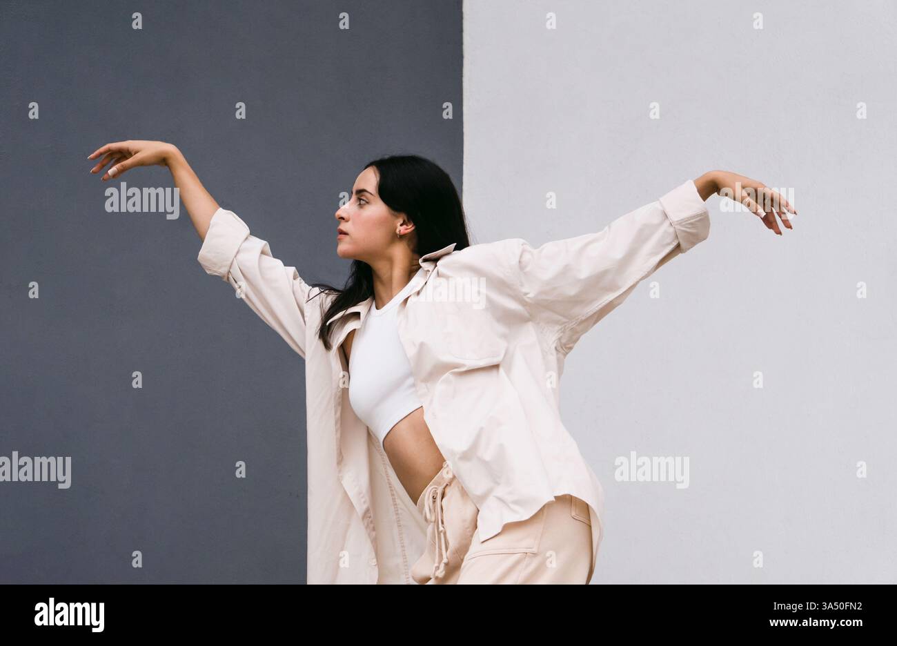 Hispanische Tänzerin mit Armen gegen eine graue und weiße Wand in einer energischen Street Dance Pose. Die dynamische Choreografie vermittelt Rhythmus, Talent und urbanen Stil für Performance und Mode. Stockfoto