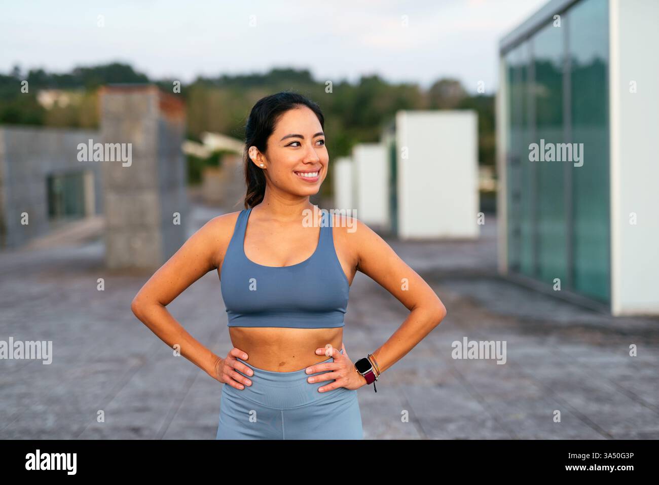 Latina Woman in Workout-Ausrüstung legt ihre Hände auf ihre Hüften und lächelt während einer Outdoor-Trainingseinheit selbstbewusst. Die Outdoor-Umgebung und die sportliche Haltung vermitteln Energie und Motivation. Perfekt für Fitness-, Wellness- und aktive Lifestyle-Kampagnen. Stockfoto