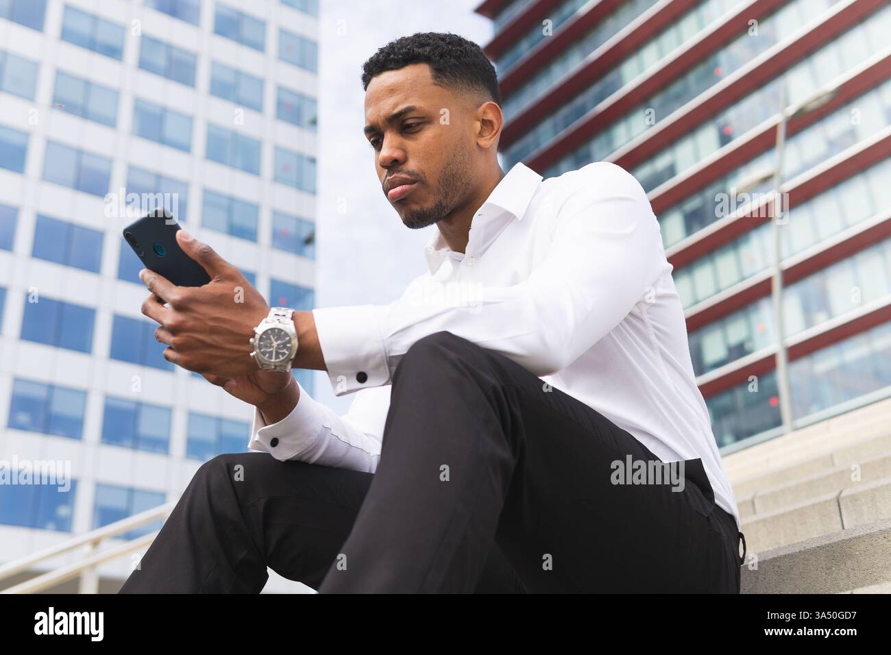 Aus einem tiefen Winkel spricht ein seriöser afroamerikanischer Mann in formeller Kleidung am Telefon, während er auf Steintreppen in einer städtischen Umgebung sitzt. Das Bild vermittelt Geschäftskommunikation und Lebensstil in der Innenstadt. Perfekt für Berufs-, Karriere- oder Arbeitsplatzkontexte. Stockfoto