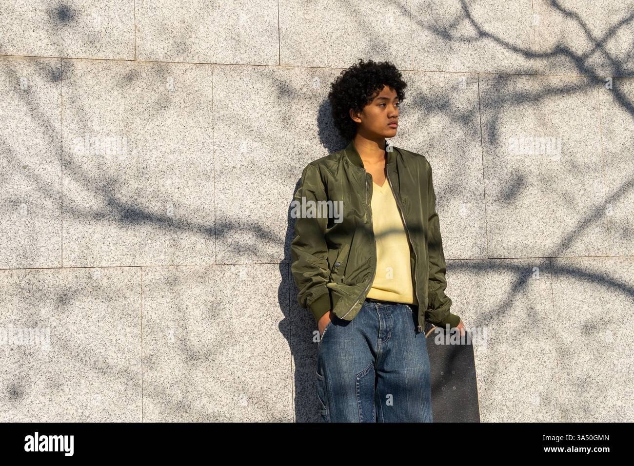 Asiatischer Mann mit Hand in der Tasche steht tagsüber mit einem Skateboard vor einer gekachelten Stadtmauer. Der ungezwungene Straßenstil vermittelt Jugendkultur und Stadtleben. Ideal für urbanes Wohnen, Street-Style und Skateboarden. Stockfoto