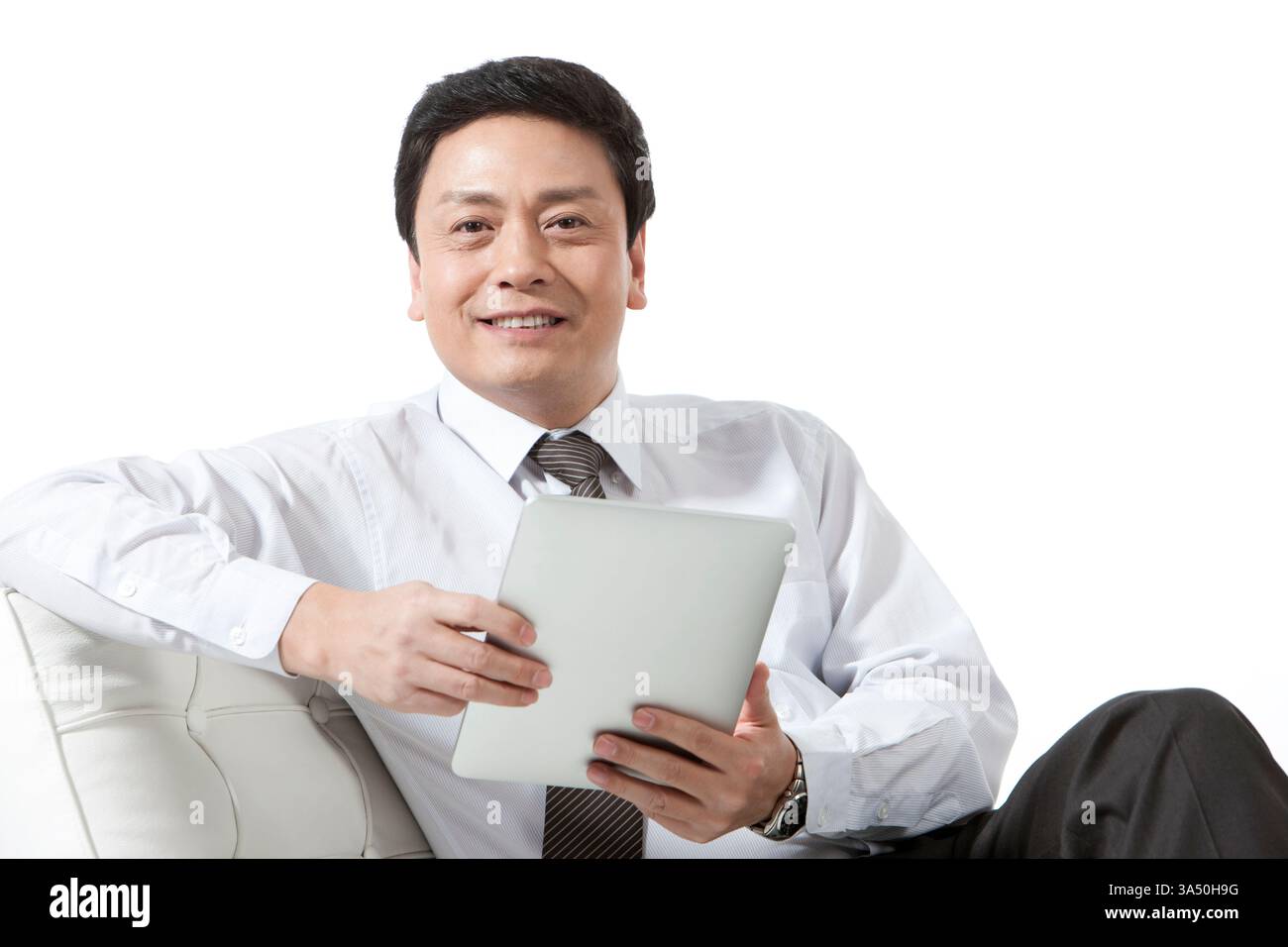 Fröhlicher chinesischer Geschäftsmann, der sich auf einem Sofa entspannt, während er ein digitales Tablet verwendet. Ein Studio mit sauberem weißem Hintergrund und modernem, professionellem Flair, ideal für Technologie, Business und Unternehmenskommunikation. Stockfoto