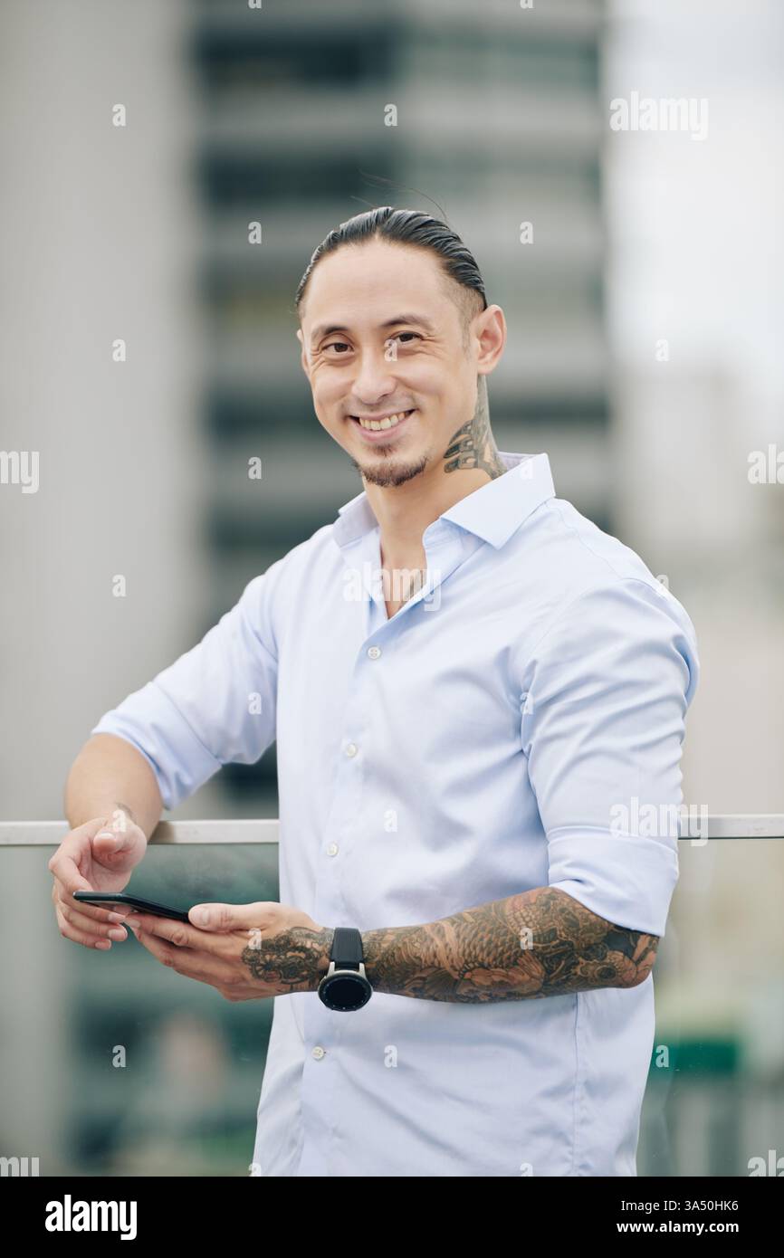 Fröhlicher Mischling-Mann auf dem Dach, lächelt in die Kamera, während er auf seinem Smartphone schreibt. Dieses städtische Porträt eignet sich hervorragend für Business-, Technologie- und professionelle Lifestyle-Kampagnen. Die moderne Dachterrasse vermittelt Konnektivität und Vertrauen. Stockfoto