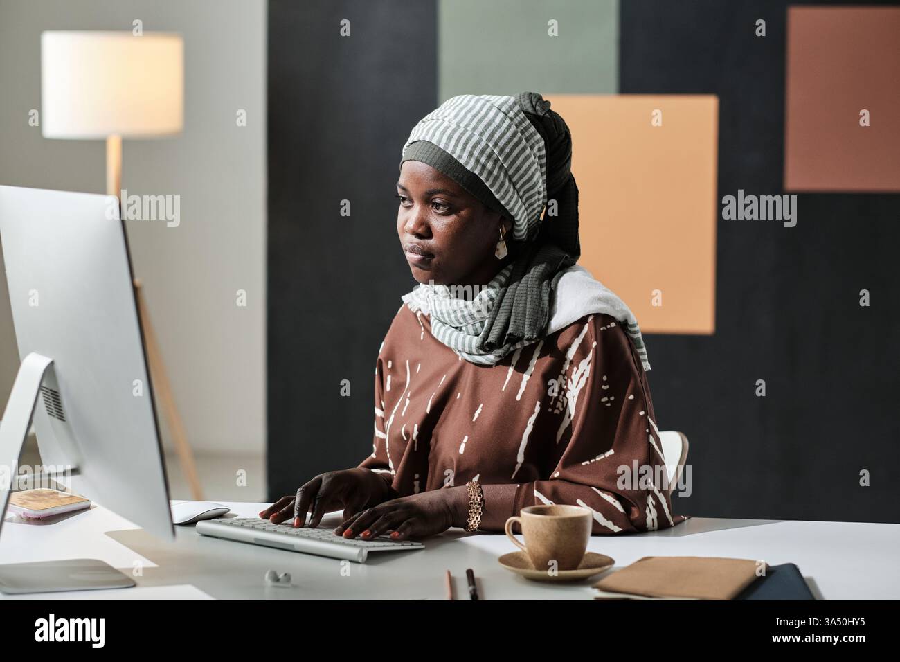 Afrikanische muslimische Geschäftsfrau, die an ihrem Schreibtisch arbeitet und am Computer schreibt. Dieses Bild in einem modernen Büro vermittelt Professionalität, Fokus und digitale Produktivität für Geschäfts- oder Unternehmergeist. Stockfoto