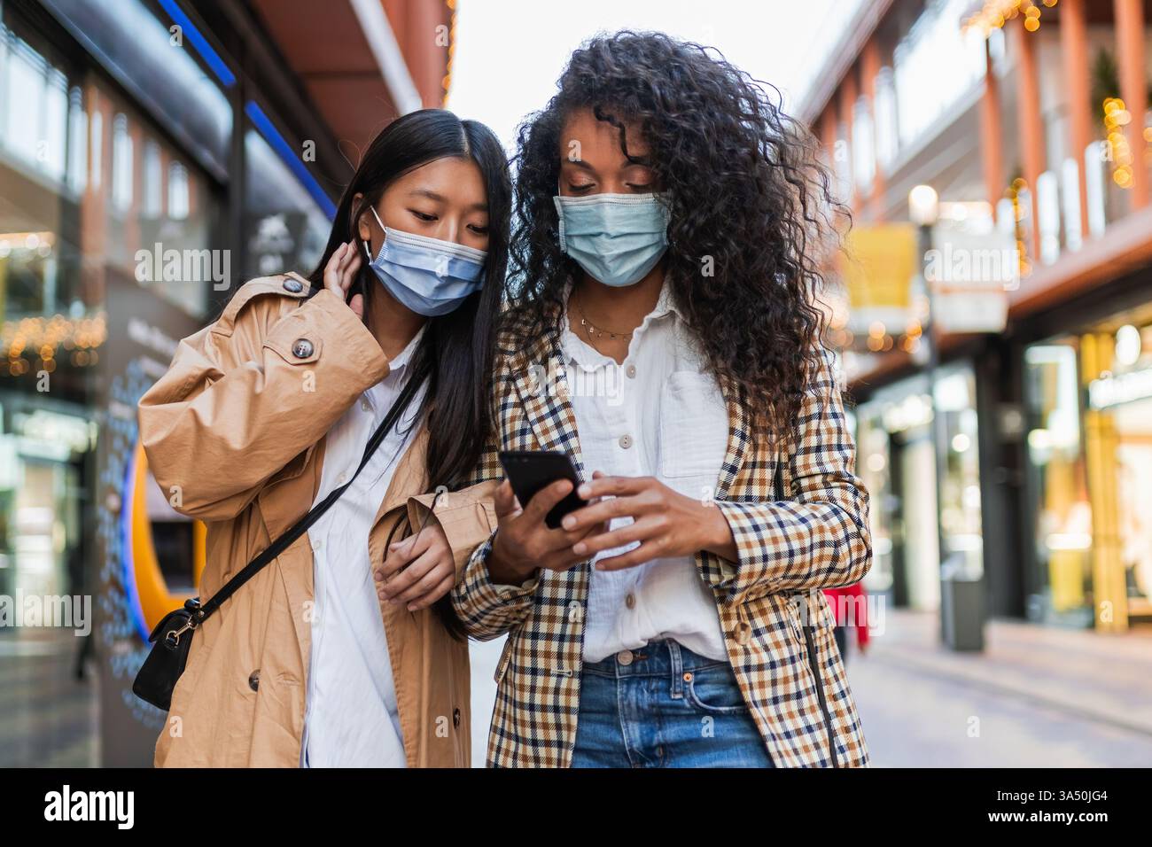 Verschiedene Frauengruppen, die Masken tragen und ein Handy auf einer Stadtstraße benutzen. Inhalte für urbanen Lebensstil und Konnektivität der COVID-19-Ära, die Vielfalt und das Straßenleben hervorheben. Stockfoto