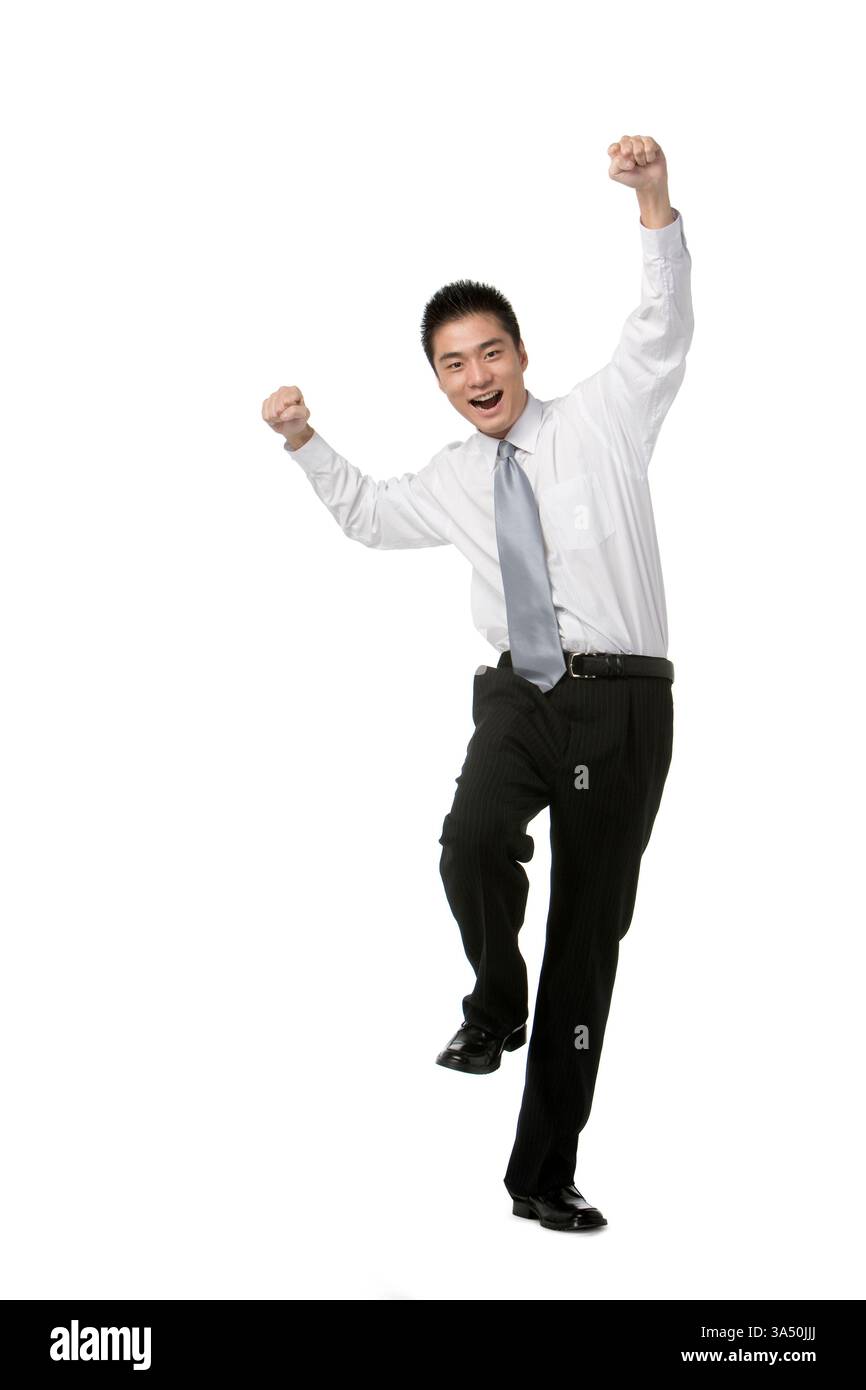 Ein fröhlicher chinesischer Geschäftsmann im Studio-Hintergrund zeigt Fäuste in einer selbstbewussten Pose mit angehobenem Bein. Dieses Image vermittelt Feier, Leistung und positive geschäftliche Dynamik. Stockfoto