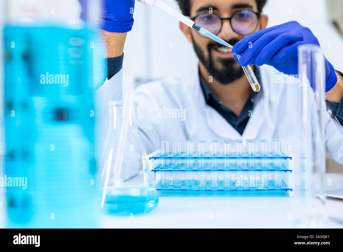 Bärtiger Wissenschaftler in Brillen und Handschuhen testet eine chemische Probe in einem Kolben im Labor. Der Innenarbeitsplatz verfügt über Glaswaren und Laborgeräte, ideal für wissenschaftliche, chemische und Forschungsthemen. Stockfoto