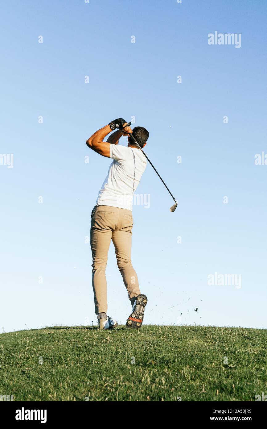 Ein männlicher Hispanic-Golfspieler steht nach seinem Schwung auf einem üppig grünen Golfplatz und blickt weg. Die sonnige Outdoor-Szene bietet Golfausrüstung und ein gut gepflegtes Fairway. Perfekt für Sport, Freizeit und Outdoor-Lifestyle. Stockfoto
