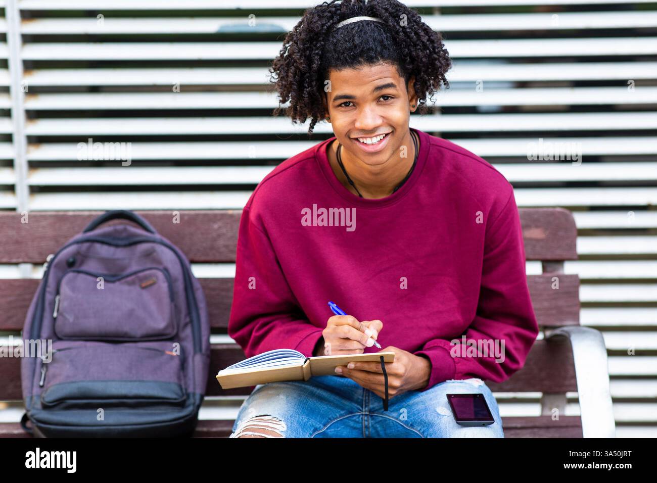 Ein junger Afroamerikaner sitzt draußen und lächelt, während er in ein Notizbuch schreibt. Die Szene zeigt Lernen, Jugend und Outdoor-Lernen in einer ungezwungenen Umgebung. Perfekt für Bildung, Studentenleben und Lifestyle-Inhalte. Stockfoto