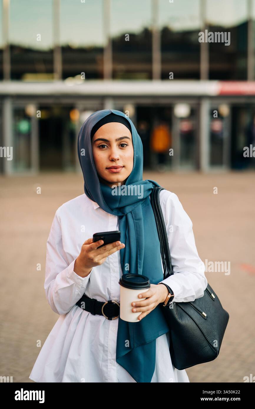 Frau aus dem Nahen Osten in einem Hijab, die ein Smartphone und einen Drink zum Mitnehmen in einer städtischen Umgebung hält. Ein zeitgenössisches Lifestyle-Image, das sich für Technologie-, Reise- und Städtekampagnen eignet. Stockfoto