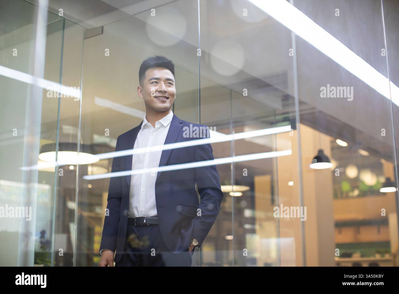 Lächelnder chinesischer Geschäftsmann, der hinter einer Glaswand im Büro wegschaut Stockfoto
