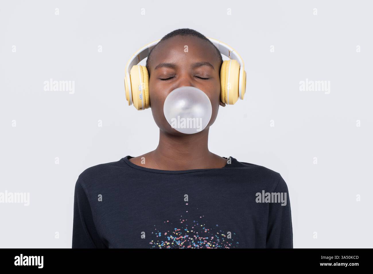 Die zufriedene junge Schwarze Frau hört Musik mit Kopfhörern, geschlossenen Augen und einer verspielten Kaugummiblase. Die saubere Studioeinrichtung unterstreicht ein fröhliches Lifestyle-Image, das sich perfekt für Musik und Mode eignet. Stockfoto