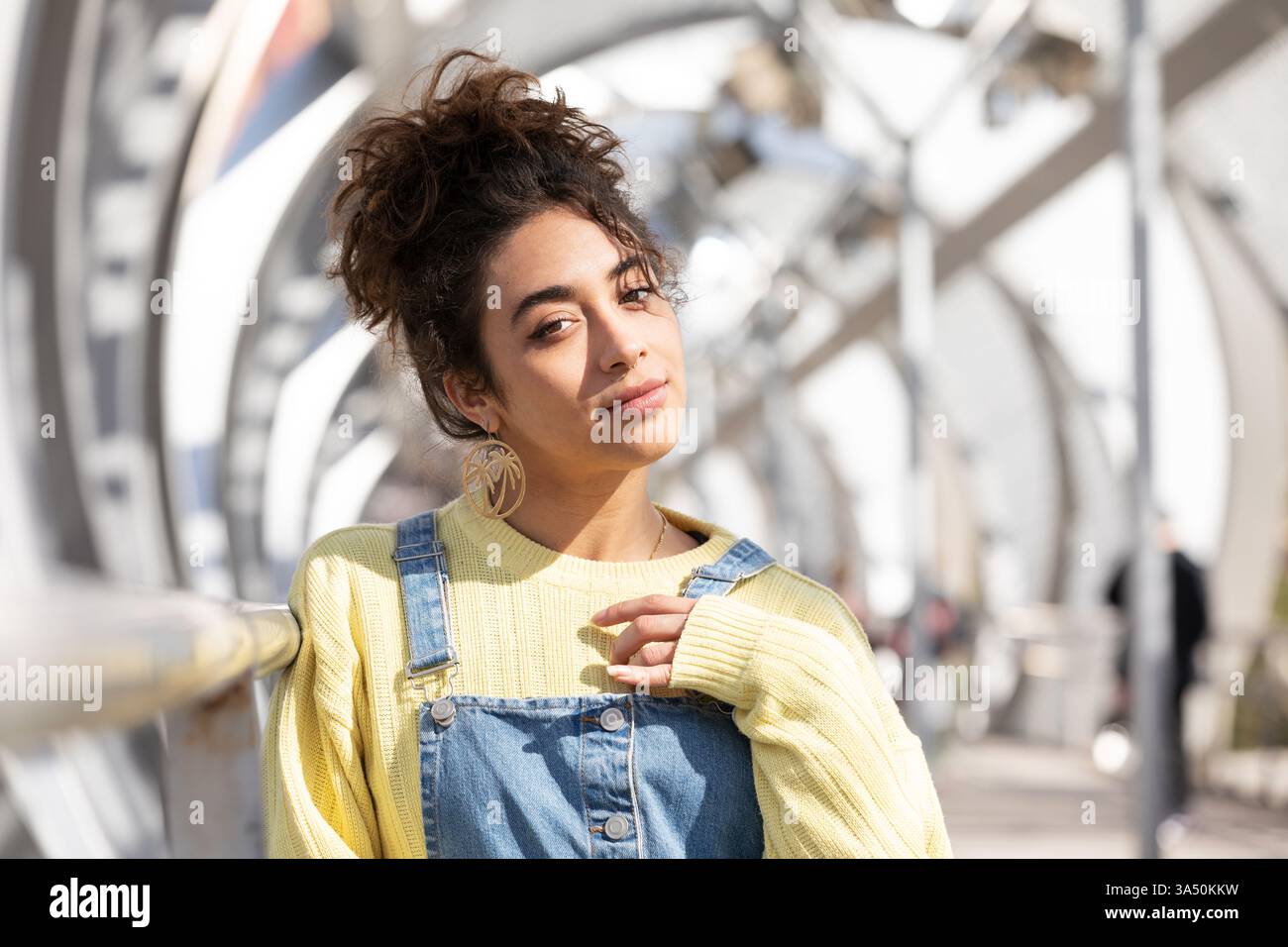 Lächelnde hispanische Frau, die sich auf einem Brückengeländer in einer sonnigen Stadt lehnt. Lässiger urbaner Style und Outdoor-Lifestyle sind perfekt für Reisen und Kampagnen im Stadtleben. Ideal für modebewusste, selbstbewusste Bilder des weiblichen Lebensstils. Stockfoto
