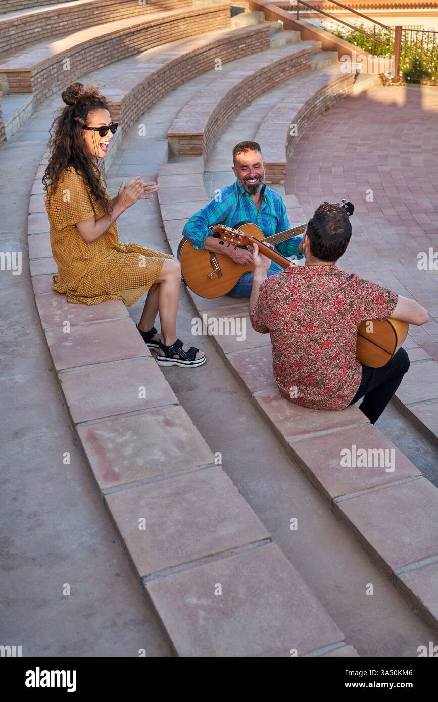 Eine Ganzkörper-Straßenszene mit drei männlichen Musikern und einer fröhlichen Frau, die Akustikgitarren im Freien auf den Stufen der Stadt spielt. Sie lächeln und sehen sich an, strahlen Freundschaft und Energie aus. Ein vielseitiges Bild für Straßenauftritte, Lifestyle im Sommer und urbane Musik. Stockfoto