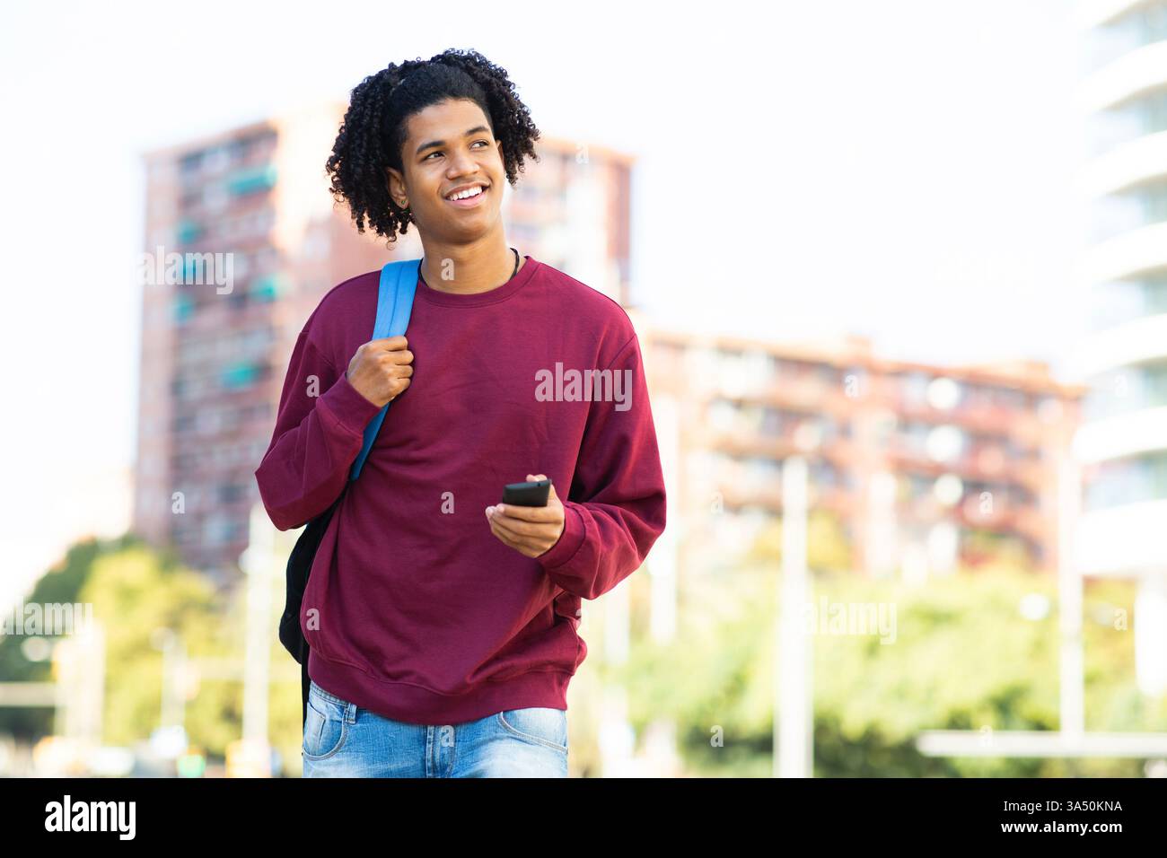 Lächelnder Mann in einer städtischen Umgebung, tagträumend mit Handy und Rucksack in der Hand. Diese lässige, fröhliche Lifestyle-Aufnahme ist ideal für Reisen, Pendeln und alltägliche technische Themen. Ideal für Kampagnen, die sich auf Jugend, Stadtleben und positives Denken konzentrieren. Stockfoto